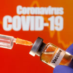 FILE PHOTO: A small bottle labeled with a "Vaccine" sticker is held near a medical syringe in front of displayed "Coronavirus COVID-19" words in this illustration
