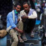 Men wearing protective face masks on their chins watch a video on a mobile phone as they sit at a second-hand motor parts market in the old quarters of Delhi