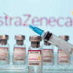 Vials labelled "AstraZeneca COVID-19 Coronavirus Vaccine" and a syringe are seen in front of a displayed AstraZeneca logo
