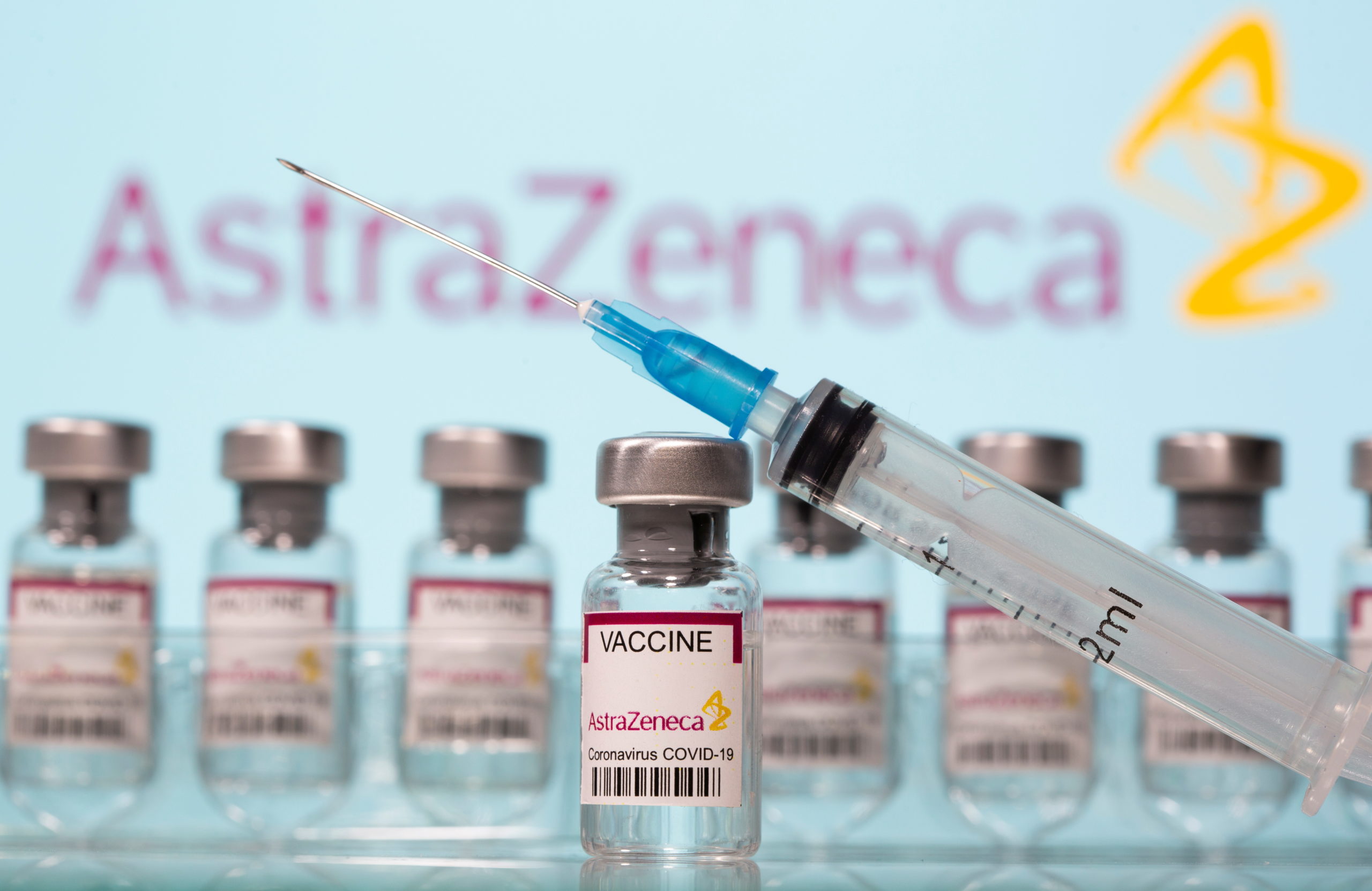 Vials labelled "AstraZeneca COVID-19 Coronavirus Vaccine" and a syringe are seen in front of a displayed AstraZeneca logo