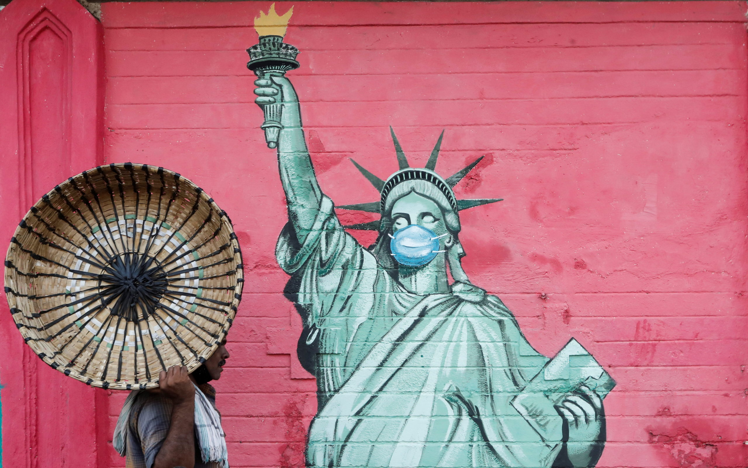Man carrying a basket walks past a graffiti depicting the Statue of Liberty wearing a mask amidst the spread of the coronavirus disease, in Mumbai