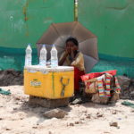 A girl selling water uses an umbrella to protect herself from the sun as she waits for customers on a hot summer day, in New Delhi