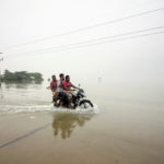 People ride on a bike on a submerged road during a widespread flood in Sylhet